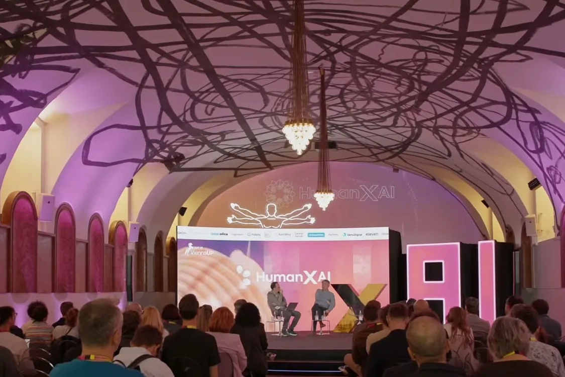 The vaulted hall at Hofstallungen MUMOK during Human × AI Europe: an audience facing the stage, where two speakers are in conversation.